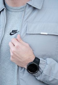 Veste matelassée gris clair avec une fermeture éclair et une poche poitrine, portée sur un t-shirt Nike gris ; montre digitale noire au poignet.