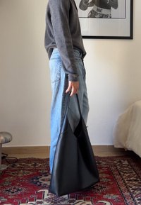 Μαύρη δερμάτινη τσάντα tote με απαλή υφή, φαρδιές ρίγες και μινιμαλιστικό σχέδιο, κρατημένη από ένα άτομο που φοράει γκρίζα ζακέτα και μπλε τζιν.