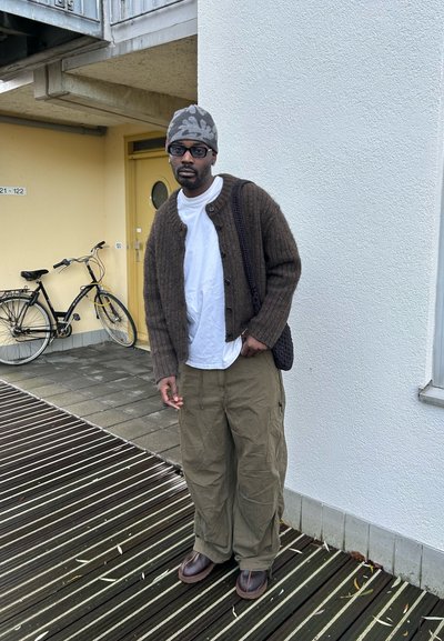 Cardigan marrón de punto sobre una camiseta blanca, combinado con pantalones khaki holgados y botas marrones. Goro gris y una bolsa negra. Bicicleta de fondo.