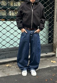 Person står med hendene i lommene iført svart hettegenser med glidelås, vide blå jeans med tre knapper, hvite joggesko, foran en metallport.