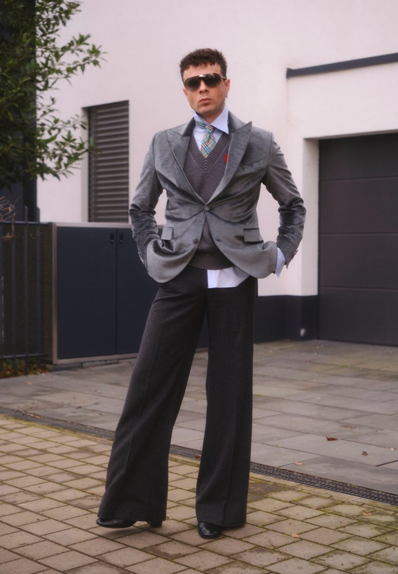 Homme portant un blazer en velours gris, un pantalon large anthracite, un gilet gris, une cravate à carreaux, des lunettes de soleil, debout sur un trottoir urbain les mains dans les poches.