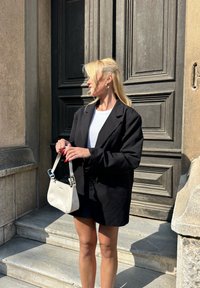 Blazer nero oversize con spalle imbottite, t-shirt bianca e borsa bianca lucida. In piedi su gradini di pietra vicino a una porta in legno testurizzata.