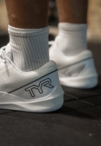 Weiße Sportschuhe mit einer strukturierten Oberfläche, ausgestattet mit einem schwarzen "TYR"-Logo und einem eleganten Design. Kombiniert mit gerippten weißen Socken.