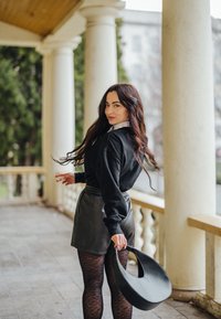 Sweatshirt noir surdimensionné, shorts en cuir et collants à motifs. Tenant un sac à main noir incurvé, debout sur un porche avec des colonnes.