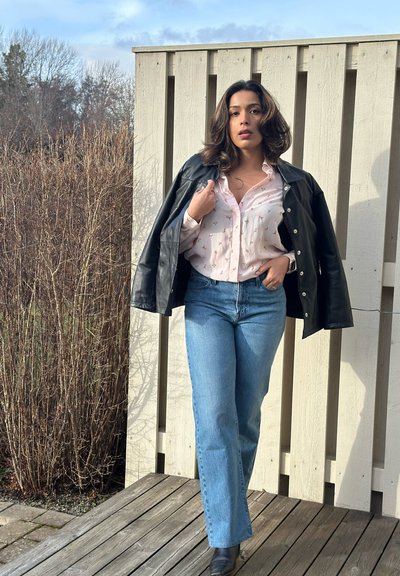 Chaqueta de cuero negra sobre una camisa de botones rosa claro con patrones florales, combinada con jeans de tiro alto de color azul y botas negras hasta el tobillo. Fondo de madera.