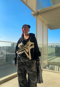 Camisola preta com fecho zip e gráfico de estrela branca, combinada com calças cinzas de camuflagem. Boné preto e bolsa com tachas completam o look. Céu brilhante ao fundo.