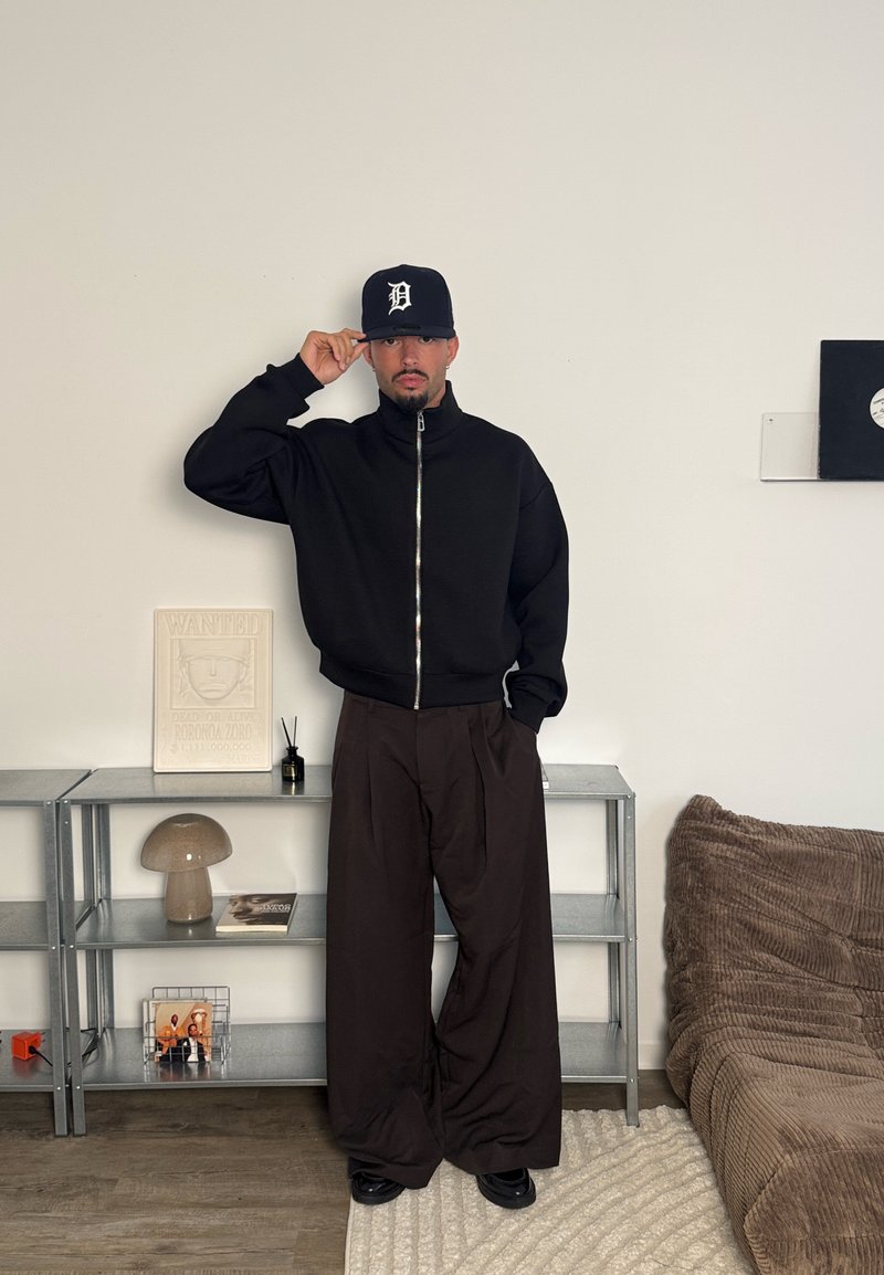 Veste noire zippée avec col montant ; pantalon marron à jambes larges ; casquette noire avec logo blanc ; debout devant un fond neutre.
