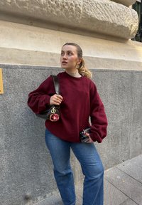 Sweatshirt oversized em borgonha com ombros caídos, combinado com jeans azuis. Segura uma carteira preta com um porta-chaves vermelho e uma câmara.