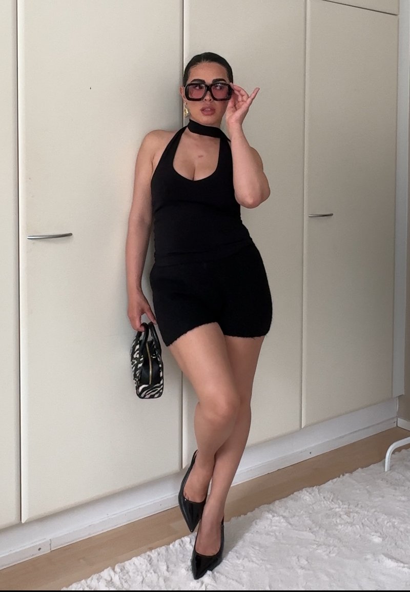 Femme posant à l'intérieur en haut halter noir, short noir, talons hauts noirs, grandes lunettes de soleil et tenant un petit sac à main à motif zèbre.