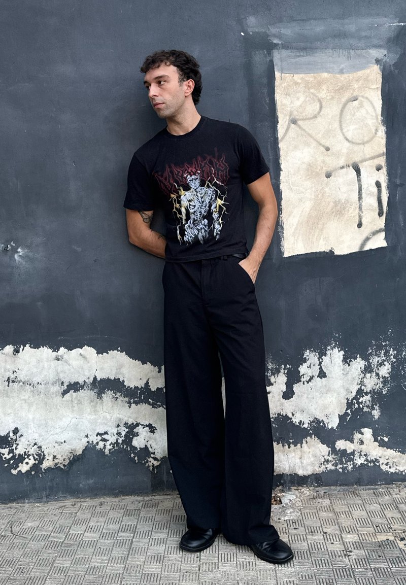 T-shirt grafico nero con design di scheletro e elementi di fulmine, abbinato a pantaloni neri larghi e scarpe nere su uno sfondo di muro texturizzato.