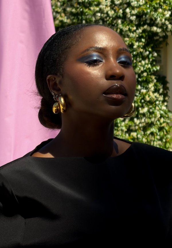 Mujer con piel oscura, sombra de ojos azul, labios oscuros y brillantes, pendientes de aro dorados, vistiendo una camiseta negra, parada al aire libre con fondos rosa y verde.