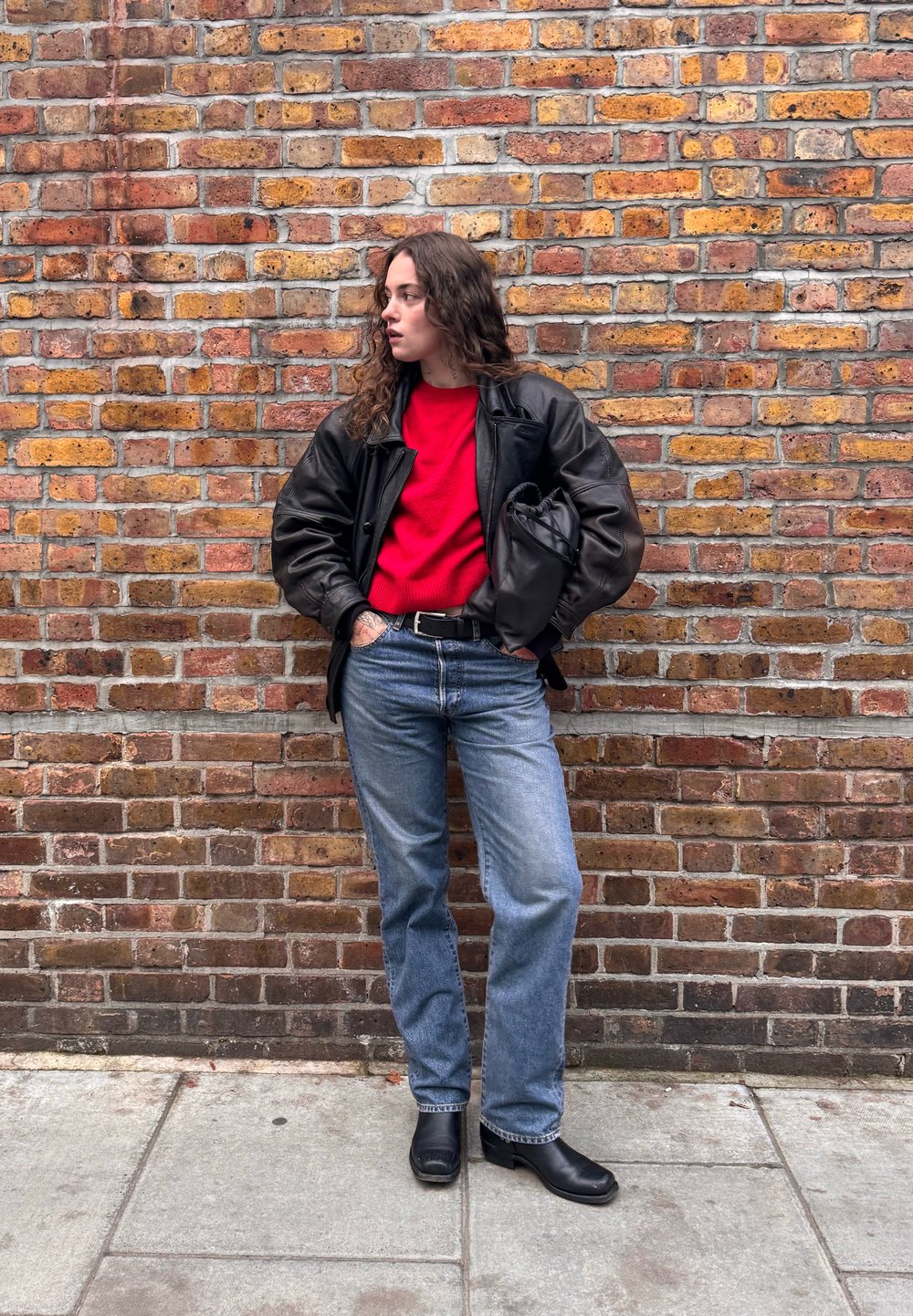 Giacca di pelle nera, maglione rosso, jeans blu, stivaletti neri alla caviglia. In piedi contro un muro di mattoni lavorato, mani in tasca, posa rilassata.