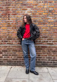 Giacca di pelle nera, maglione rosso, jeans blu, stivaletti neri alla caviglia. In piedi contro un muro di mattoni lavorato, mani in tasca, posa rilassata.