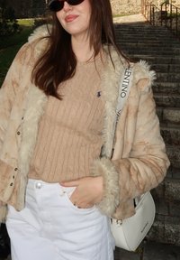 Chaqueta beige con forro de piel, suéter de punto con diseño de cables debajo, jeans blancos y un bolso blanco. Gafas de sol completan el conjunto.