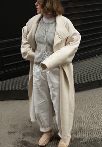 Femme portant un long manteau beige, un cardigan gris, un pantalon blanc et des chaussures beiges, debout sur un trottoir urbain, regardant vers la gauche.