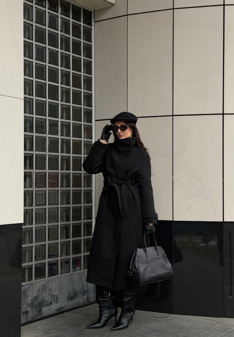 Black wool overcoat with a belt, black gloves, and knee-high stiletto boots. Accessories include a black handbag and a flat cap.