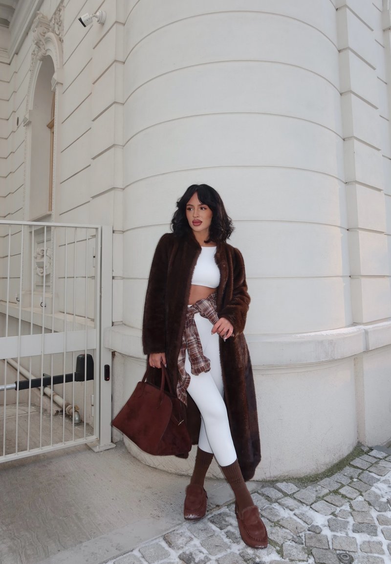 Vrouw die een donkerbruine lange jas draagt, een wit crop top en legging, bruine sokken en schoenen, en een grote bruine tas vasthoudt bij een wit gebouw.