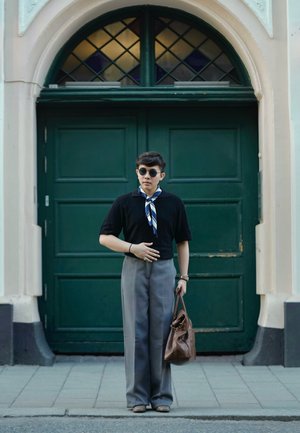 Camisa de punto negra de manga corta con una bufanda a rayas, pantalones grises de cintura alta y un bolso de cuero marrón, de pie frente a unas puertas dobles verdes.