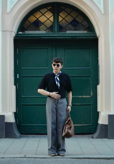 Camisa de punto negra de manga corta con una bufanda a rayas, pantalones grises de cintura alta y un bolso de cuero marrón, de pie frente a unas puertas dobles verdes.