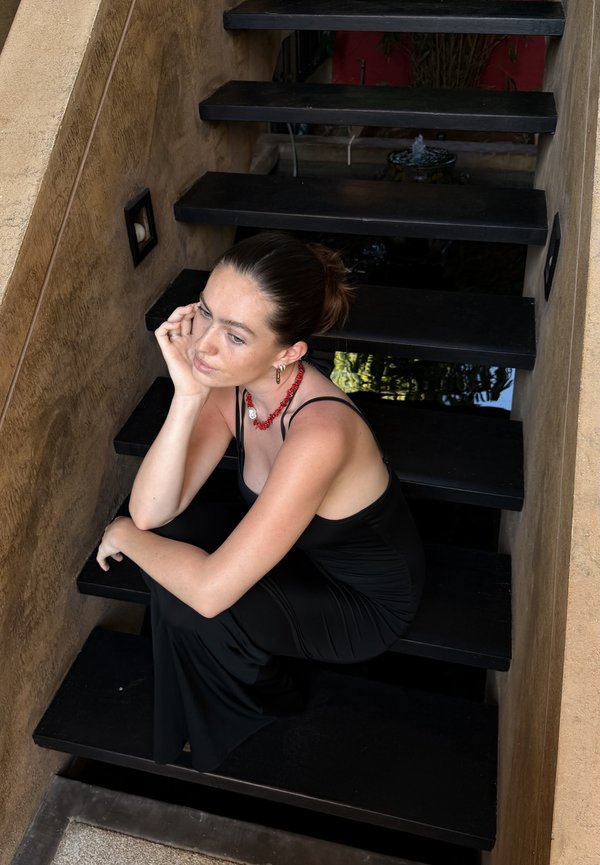 Jauna moteris juodoje suknelėje ir raudonu vėriniu sėdi apmąstanti ant juodų laiptų prie gelsvų sienų, galvą atsiremusi į vieną ranką.