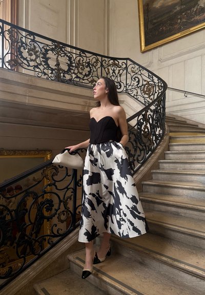 Top negro sin tirantes combinado con una falda blanca fluida que presenta un atrevido patrón abstracto en negro; complementado con un bolso crema y tacones negros.