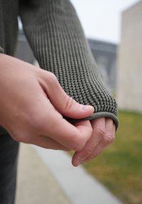 Poignet de pull vert olive texturé tenu par une main, présentant un motif côtelé et un ajustement serré autour du poignet. En arrière-plan, de l'herbe floue.