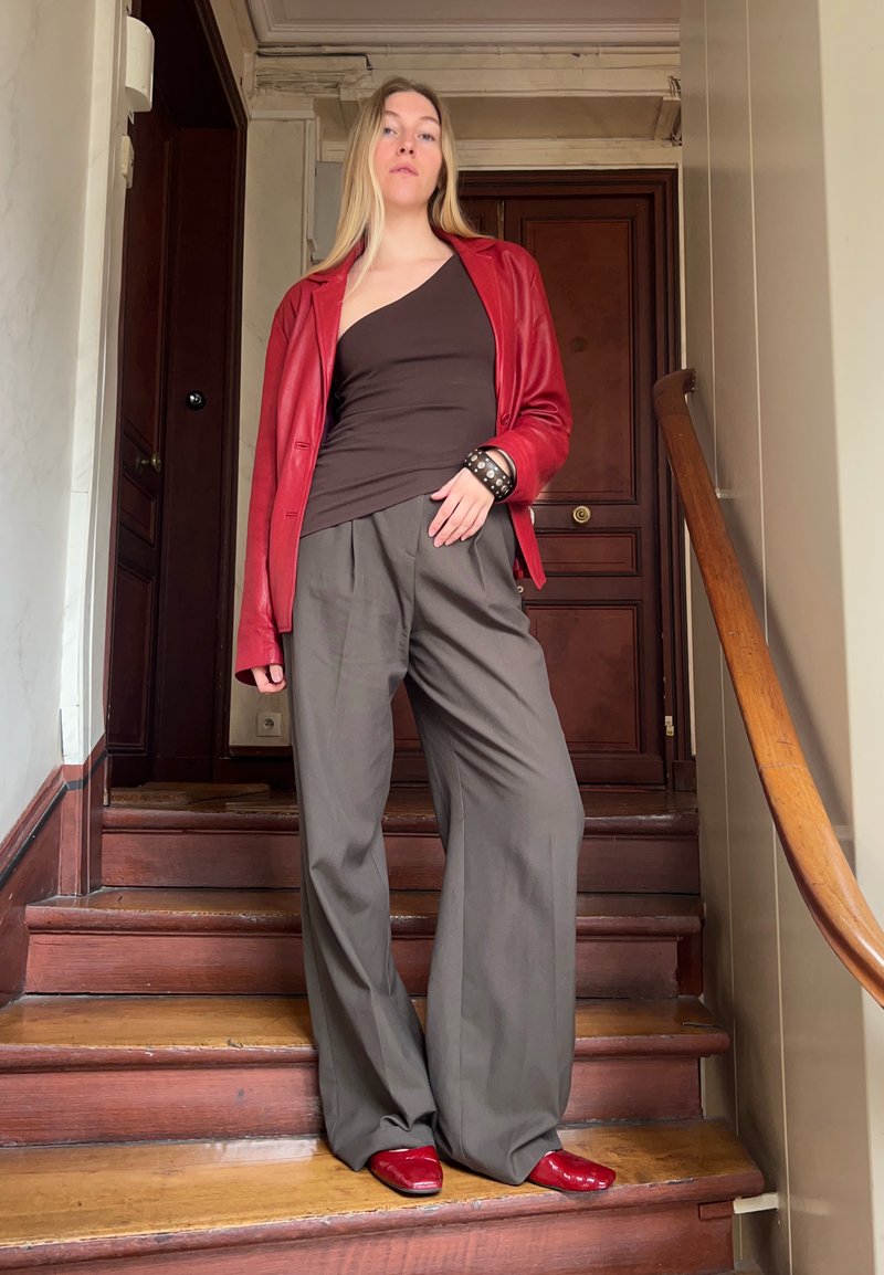 Red leather blazer, brown one-shoulder top, grey wide-leg trousers, and shiny red shoes. Wooden stairs in a well-lit indoor setting.