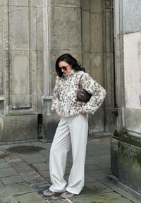 Ψεύτικο γούνινο μπουφάν σε ανοιχτό μπεζ χρώμα με καφέ στίγματα, συνδυασμένο με λευκά παντελόνια με φαρδιά πόδια. Καφέ τσάντα με μεταλλικά στοιχεία, γυαλιά ηλίου και αθλητικά παπούτσια.