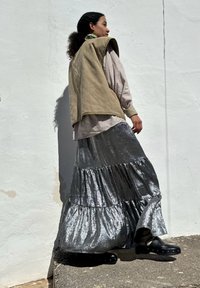 Asymmetrisk beige väst över en mönstrad skjorta, kombinerad med en lagrad, metallisk silverkjol och svarta träskor, mot en vit vägg.