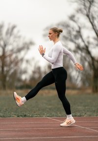 Idrottare i vit långärmad tröja och svarta leggings, springer på en bana. Bär en klocka, skorna har en ljus persikofärg med accenter.