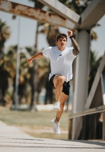 Joven con tatuajes que lleva una camisa blanca y pantalones cortos negros saltando al aire libre cerca de una estructura metálica y palmeras.