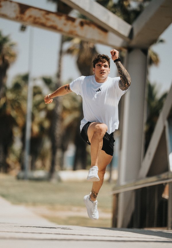 Joven con tatuajes que lleva una camisa blanca y pantalones cortos negros saltando al aire libre cerca de una estructura metálica y palmeras.
