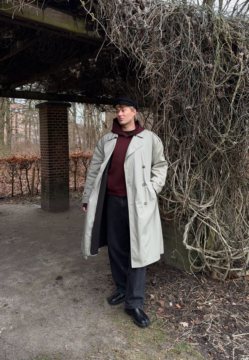 Jeune homme portant un trench-coat beige et une casquette noire, debout sous une pergola en bois recouverte de vignes nues enchevêtrées dans un parc d'hiver.