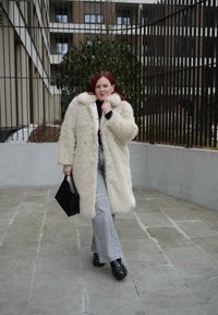 Γυναίκα με κόκκινα μαλλιά που φοράει ένα μακρύ μπεζ γούνινο παλτό, ανοιχτά γκρι τζιν, μαύρα παπούτσια με καρφιά και κρατάει μια μαύρη τσάντα στην ύπαιθρο.