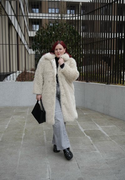 Mujer con cabello rojo que lleva un abrigo largo de piel beige, jeans gris claro, zapatos negros con tachuelas y un bolso negro al aire libre.