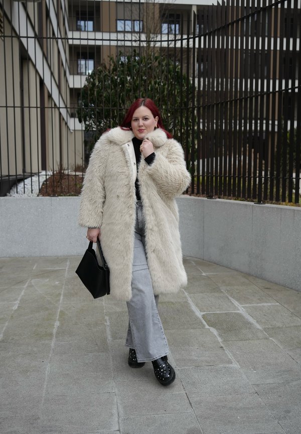 Γυναίκα με κόκκινα μαλλιά που φοράει ένα μακρύ μπεζ γούνινο παλτό, ανοιχτά γκρι τζιν, μαύρα παπούτσια με καρφιά και κρατάει μια μαύρη τσάντα στην ύπαιθρο.