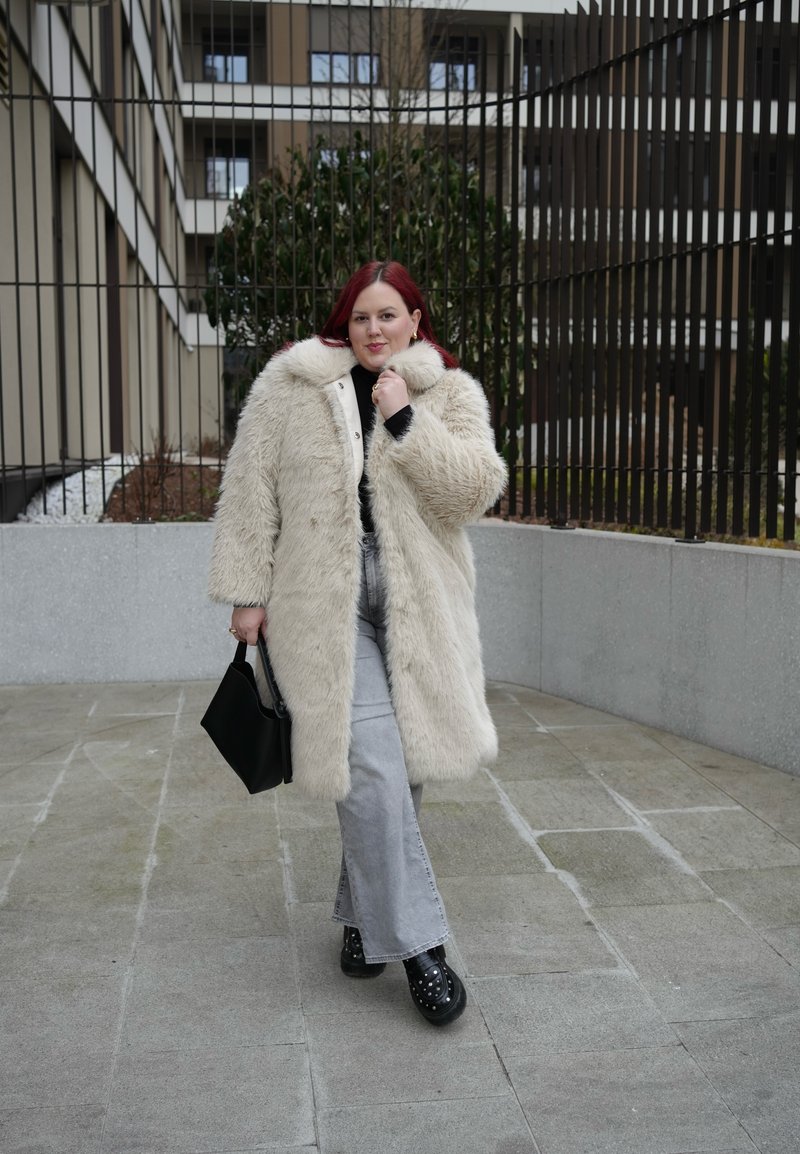 Ženska rdečih las, oblečena v dolg bež krznen plašč, svetlo sive kavbojke, črne čevlje z zakovicami ter nosi črno ročno torbico na prostem.