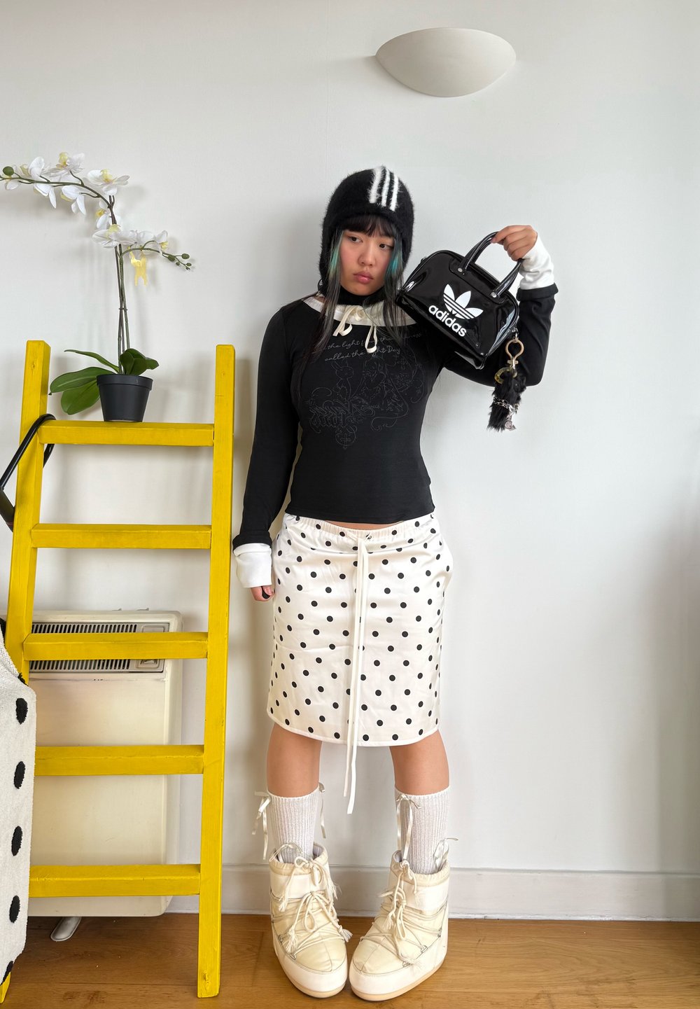 Čierny dlhý rukáv s bielymi akcentmi, sukňa s bodkami, biele podkolienky a krémové čižmy. Drží malú čiernu tašku Adidas.