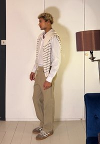 Νέος άνδρας με μπεζ παντελόνι, λευκό πουκάμισο και ριγωτό πουλόβερ περασμένο στους ώμους, στέκεται σε εσωτερικό χώρο κοντά σε καφέ λάμπα και μπλε καρέκλα.