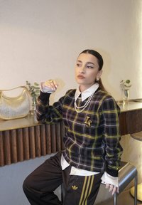 Νεαρή γυναίκα καθισμένη σε σκαμπό, φοράει πουλόβερ με καρό σχέδιο, λευκό πουκάμισο με γιακά, περιδέραιο από μαργαριτάρια και μαύρο παντελόνι Adidas με χρυσές ρίγες, δίπλα σε πάγκο.