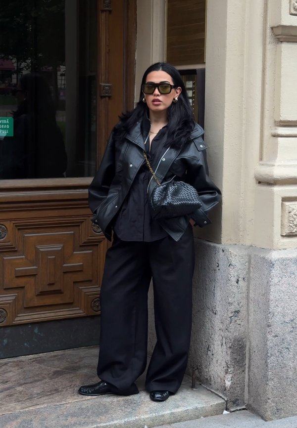 Frau trägt schwarze Lederjacke, weite schwarze Hose, Sonnenbrille und hält eine strukturierte schwarze Handtasche, steht an einer Holztür auf einem steinernen Gehweg.