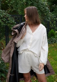 Blusa blanca a rayas con escote en V y botones, combinada con pantalones cortos a juego. Un abrigo marrón a cuadros está colgado de un brazo. Fondo verde.