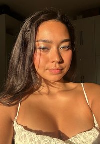 Nærbilde av en person med langt, mørkt hår som har på seg en hvit blondetopp. Myk belysning fremhever hudtonen deres og subtil sminke med blå detaljer.