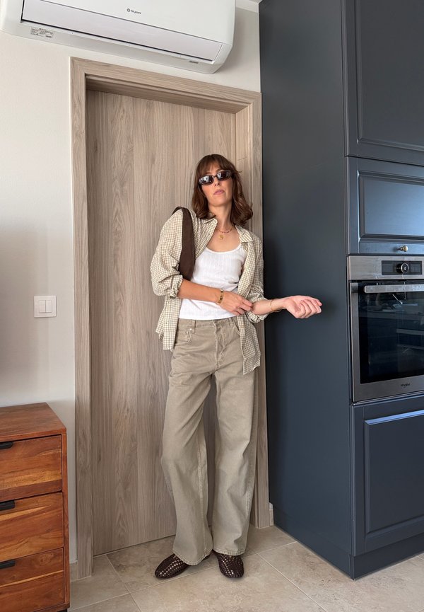 Femme portant des lunettes de soleil, une chemise beige à carreaux, un débardeur blanc et un pantalon beige ample, se tient dans une cuisine près d'une porte en bois et des armoires bleu foncé.