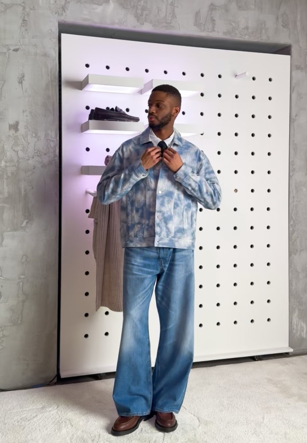 Uomo che regola la cravatta indossando una giacca blu tie-dye, jeans a gamba larga e scarpe marroni, in piedi di fronte a un pannello forato con ripiani e vestiti.