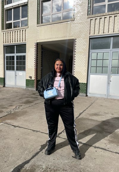 Chaqueta de cuero negra, sudadera rosa, chándal negro con rayas blancas, bolso Adidas azul, de pie sobre una superficie de concreto con edificios detrás.