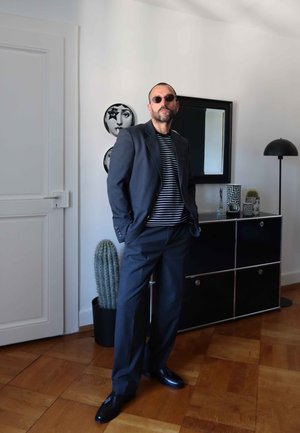 Hombre vistiendo un traje oscuro y una camisa a rayas, gafas de sol redondas. Interior moderno con un cactus, una obra de arte de espejos y un armario negro.