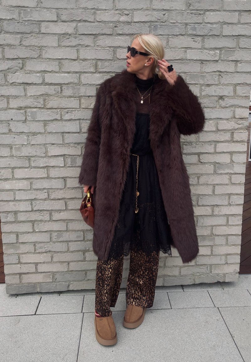 Brown faux fur coat, sheer black dress, patterned trousers, light brown footwear, and a brown handbag. Accessories include layered necklaces.