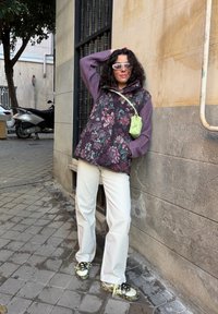 Γυναίκα με σγουρά μαλλιά, φορώντας λευκό παντελόνι, φλοράλ γιλέκο με πουπουλένια επένδυση, μοβ πουλόβερ, πράσινη τσάντα χιαστί και λευκά γυαλιά ηλίου, στέκεται σε πεζοδρόμιο της πόλης.