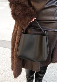 Sac seau en cuir noir à texture douce, doté d'une bandoulière unique et d'un design minimaliste. Associé à un manteau en fausse fourrure marron.